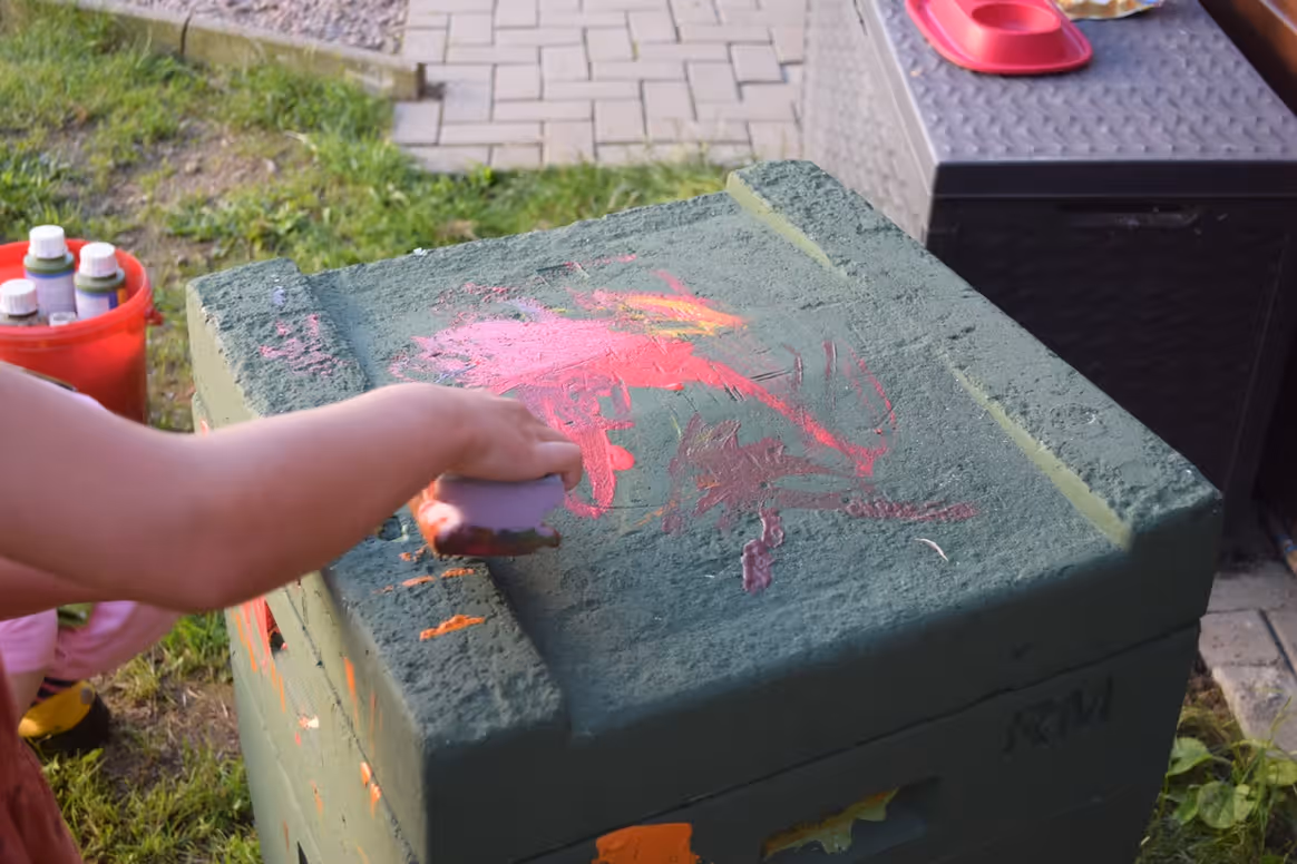 Kind malt mit einem Schwamm farbige Muster auf eine grüne, raue Oberfläche im Freien.