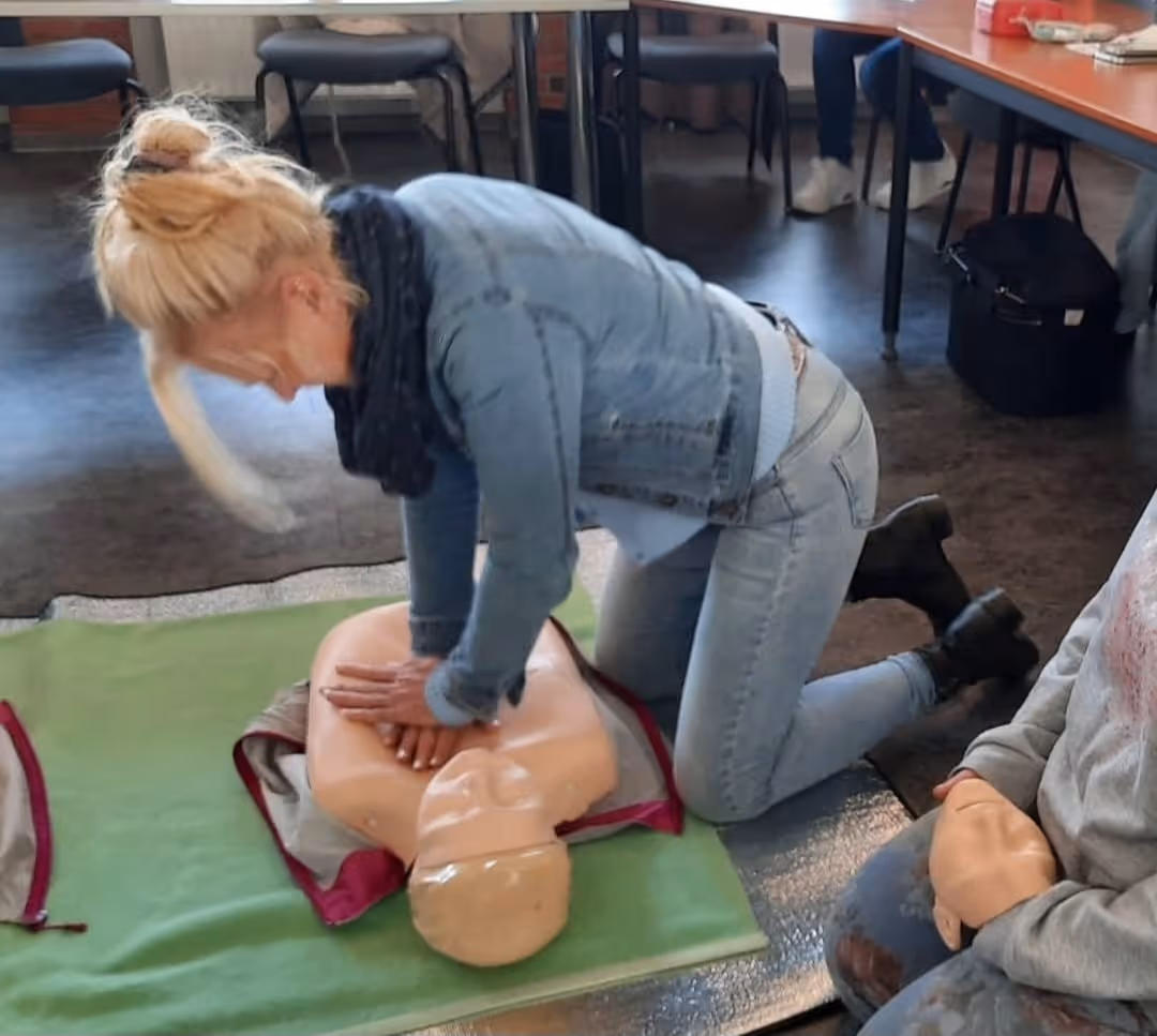 Frau in blauer Jeansjacke kniet auf Grünfläche und übt Herzdruckmassage an einer Übungspuppe.