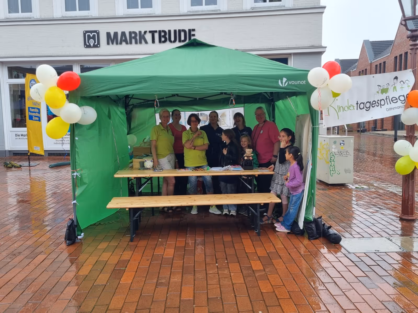 Mitglieder vom Kindertagespflege Verein, die in einem grünen Zelt mit bunten Luftballons auf einem Marktplatz stehen, neben einem Banner mit der Aufschrift 'Kindertagespflege'.