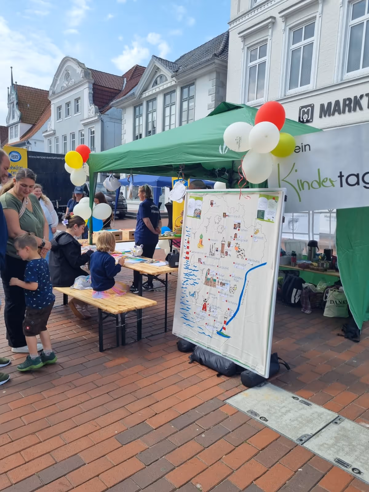Straßenfest mit einem grünen Zelt, bunter Ballon-Dekoration und Menschen, darunter Kinder, an einem Stand mit einem großen, illustrierten Poster einer Landkarte.