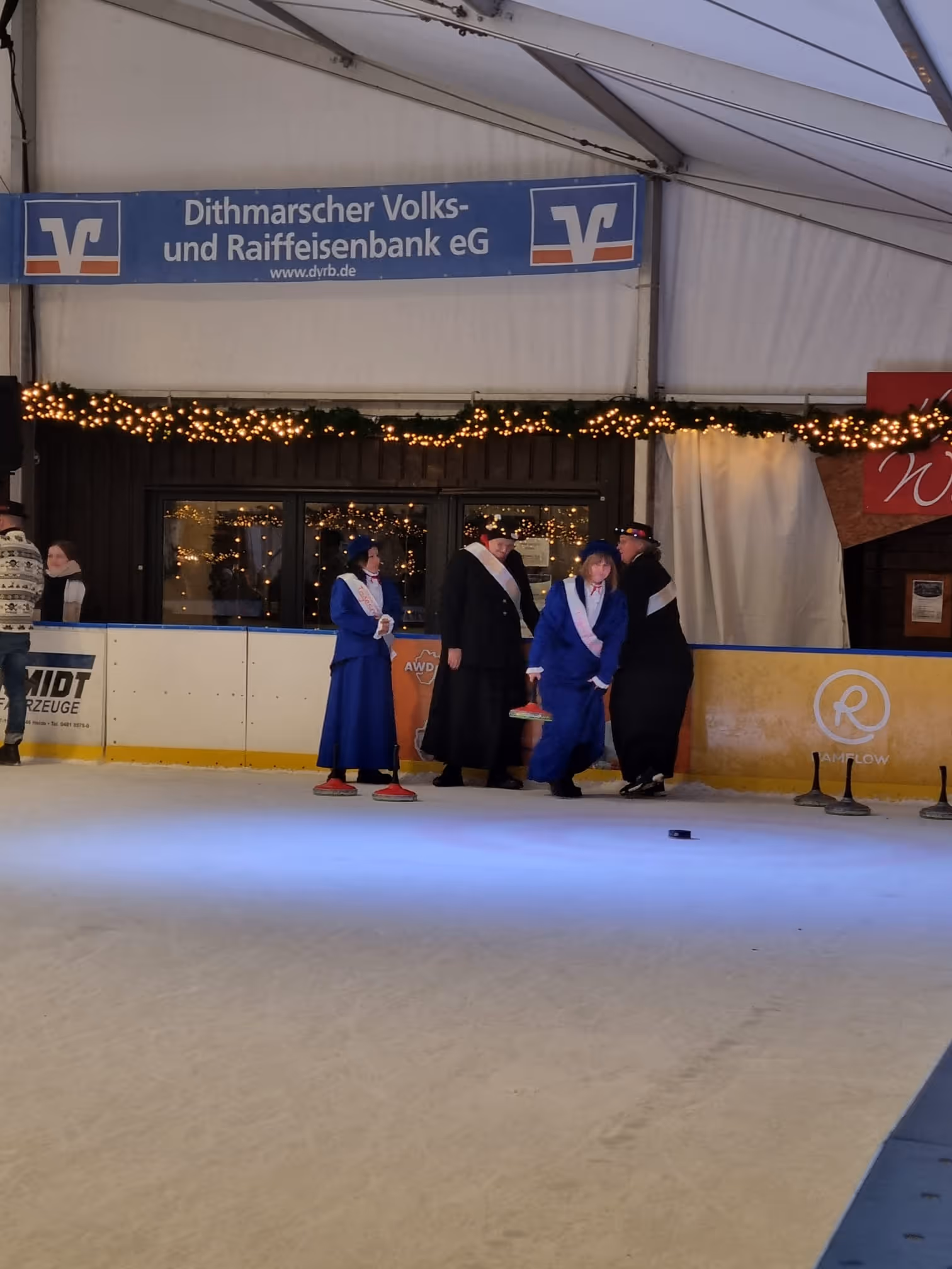Mehrere Menschen in Kostümen beim Eisstockschießen in einer überdachten Eisbahn mit Weihnachtsbeleuchtung.