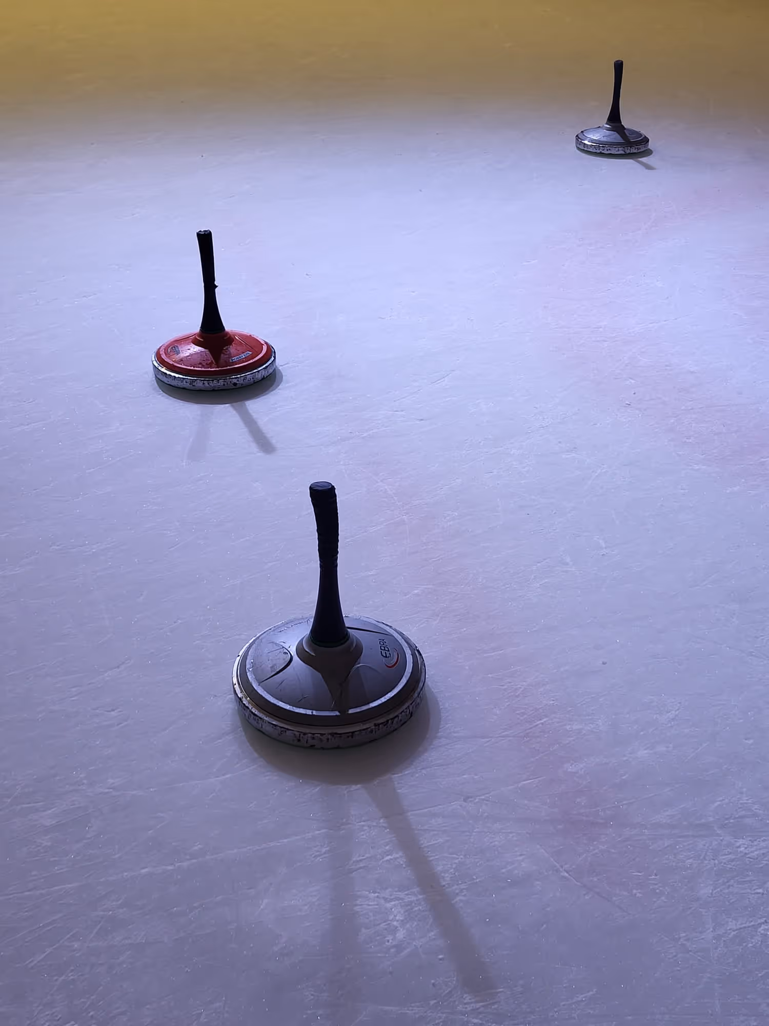 Drei Eisstockschießen auf glattem Eis, zwei in Grau und einer in Rot.