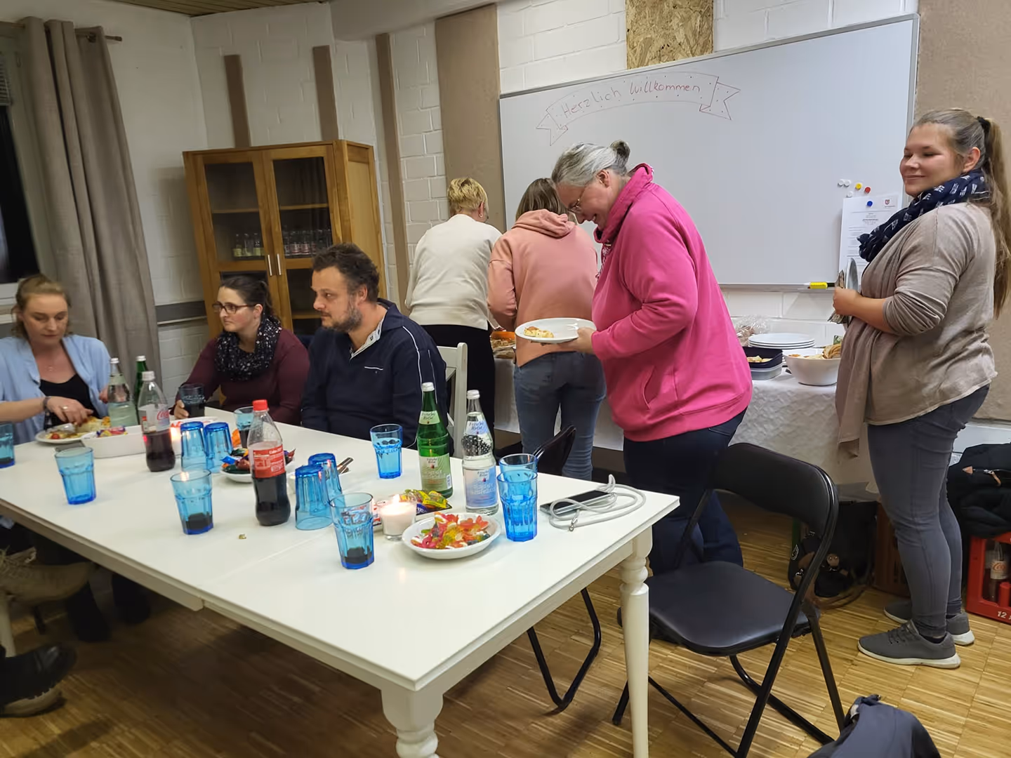 Mehrere Menschen sitzen und stehen in einem Raum mit Tischen, auf denen Getränke und Süßigkeiten stehen.