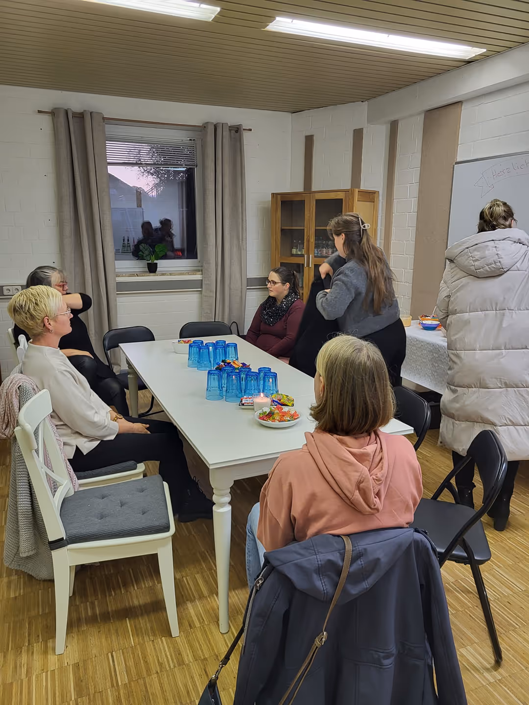 Fünf Frauen sitzen und unterhalten sich an einem weißen Tisch in einem Raum mit Holzfußboden und weißen Wänden.