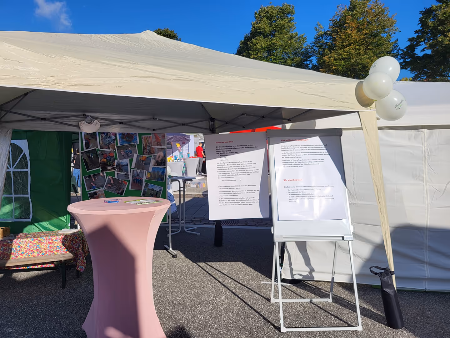 Messestand mit Informationsplakaten, rosa Stehtisch und weißen Luftballons unter weißem Pavillon.