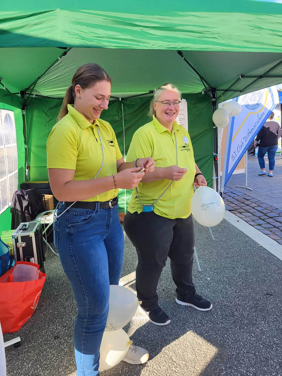 Zwei Frauen von Kindertagespflege Verein bereiten weiße Luftballons unter einem grünen Zelt vor.