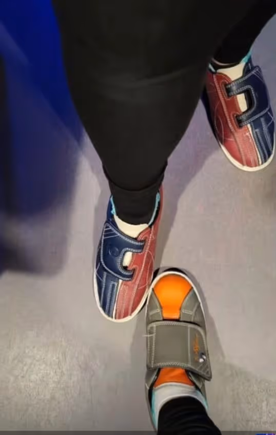 Zwei Personen tragen bunte Bowling-Schuhe, eine mit rot-blauen und die andere mit grauen und orangenen Schuhen, stehend auf einer Bowlingbahn.