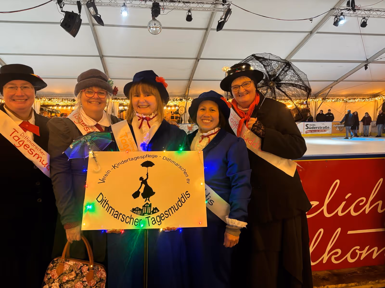 Fünf Frauen in traditionellen Kostüm mit Hütenn, die ein beleuchtetes Schild des Vereins Kindertagespflege Dithmarschen halten.
