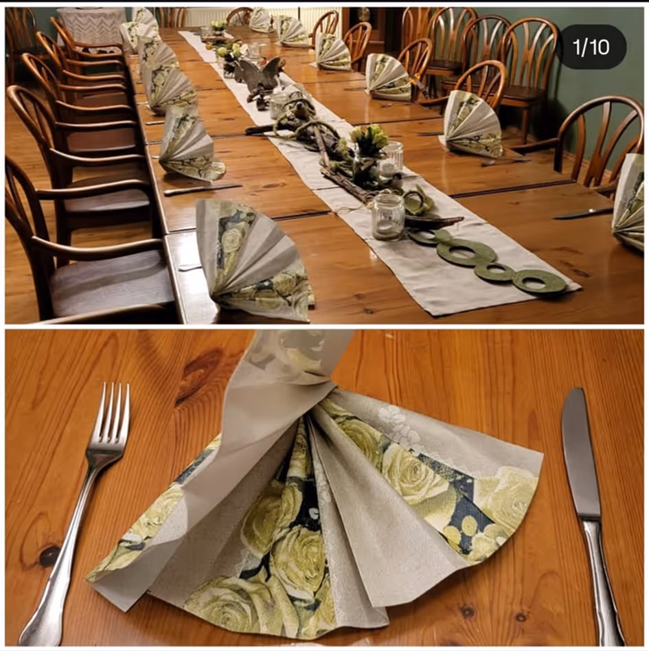 Langer Esstisch mit gefalteten Servietten in Blumenmuster, dekoriert mit Kerzen und grüner Tischdecke in der Mitte.