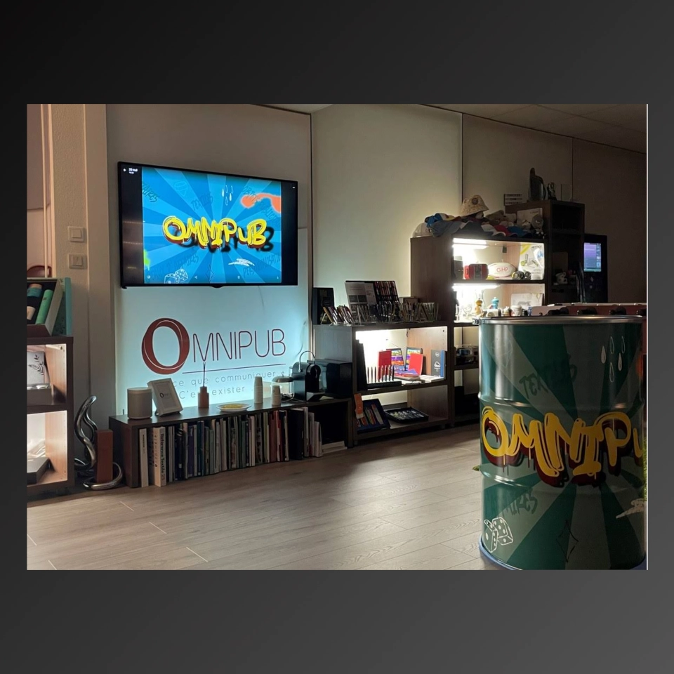 Bureau moderne avec un écran diffusant le logo Omnipub, étagères remplies de livres et fournitures, et un tonneau décoré avec le logo Omnipub.