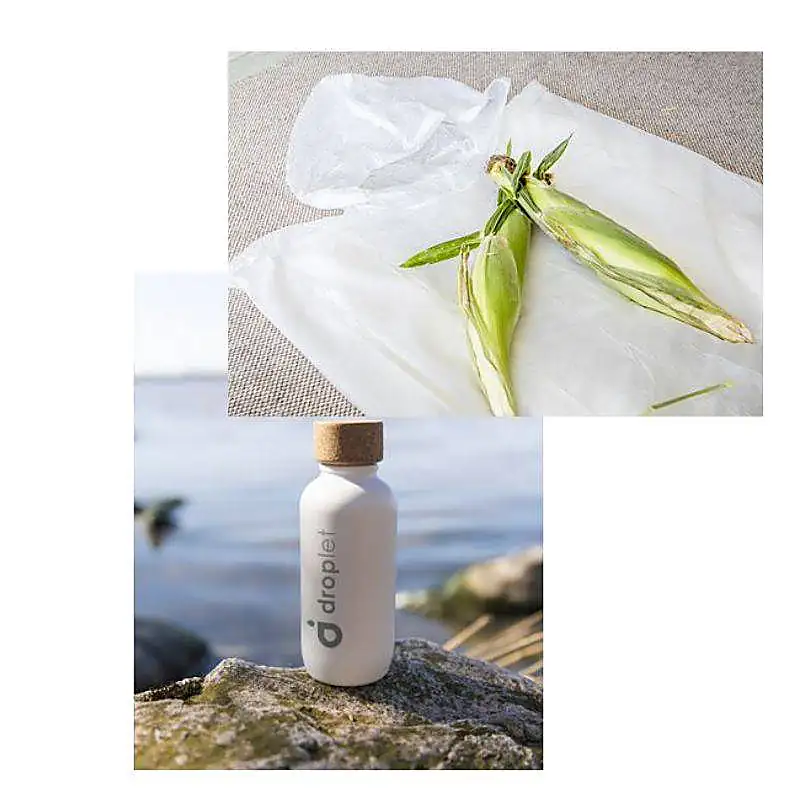 Deux épis de maïs enveloppés dans un film plastique et une bouteille blanche Droplet avec un bouchon en liège posée sur une pierre au bord de l'eau.