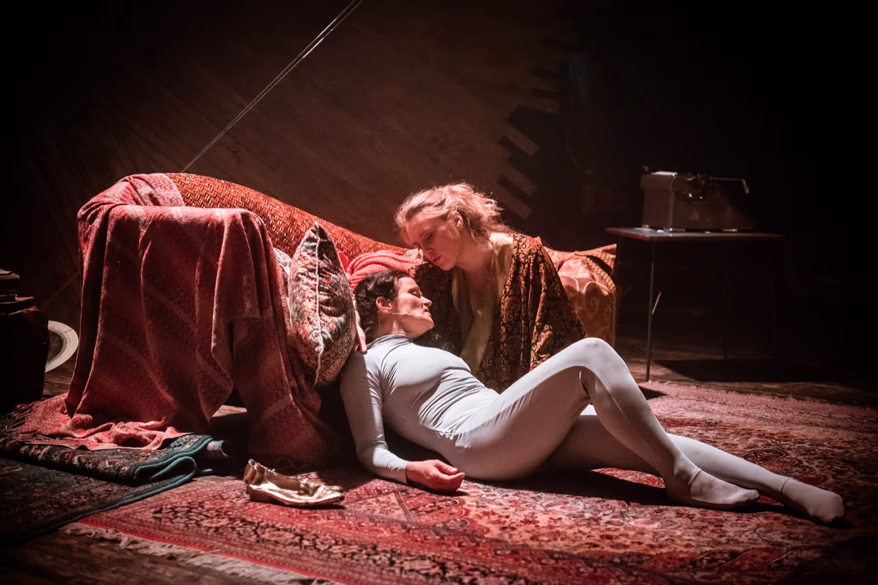 A woman in a white bodysuit lying on a patterned carpet resting her head on a red-covered sofa while another woman in a patterned robe leans over her indoors.