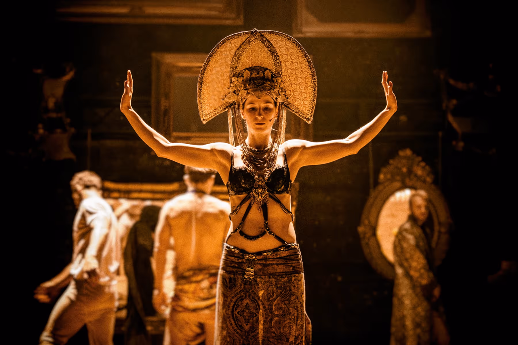 Woman dressed in elaborate costume with a large ornate headdress and patterned skirt, standing with arms raised on stage.
