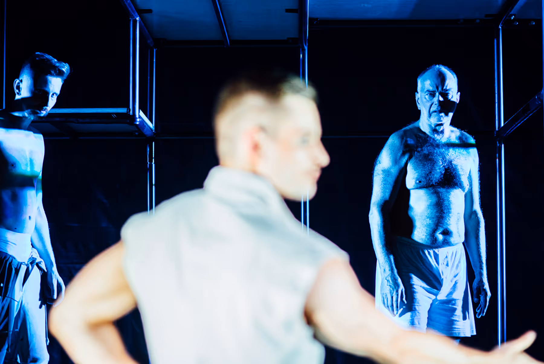 Three men in a dark room illuminated with blue light, two shirtless and one blurred in the foreground.