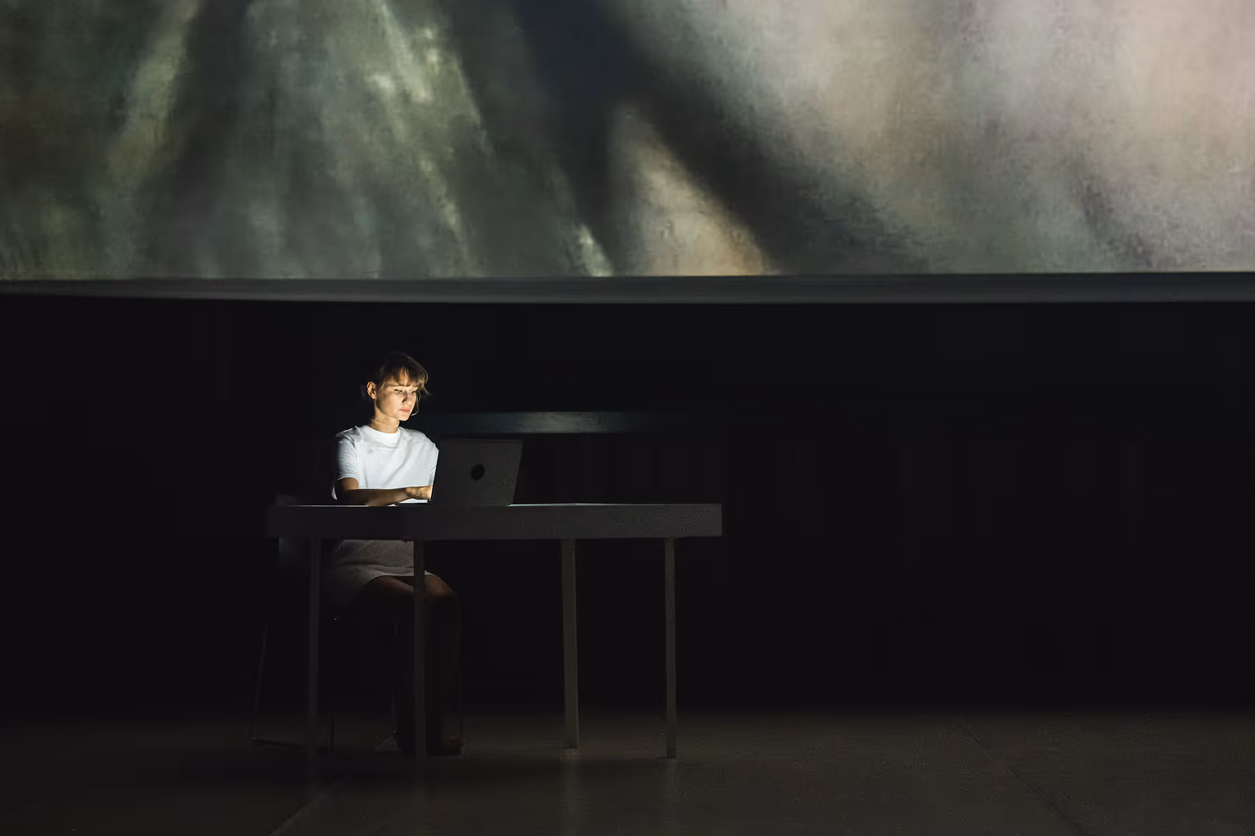 Woman in white shirt working on a laptop at a table in a dark room with a large screen behind her.