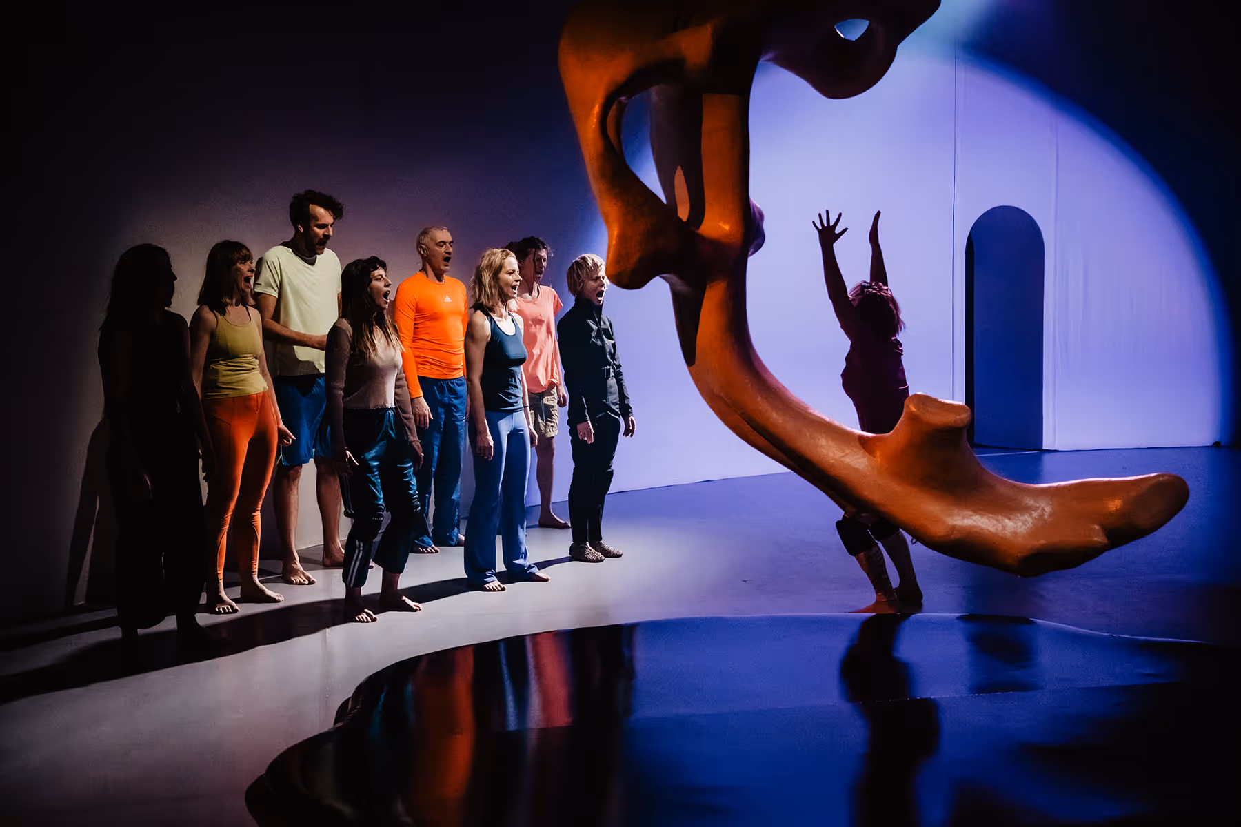 Group of people standing barefoot in a line, appearing to shout or sing towards a large orange abstract sculpture with one person reaching up behind it in a blue-lit room.