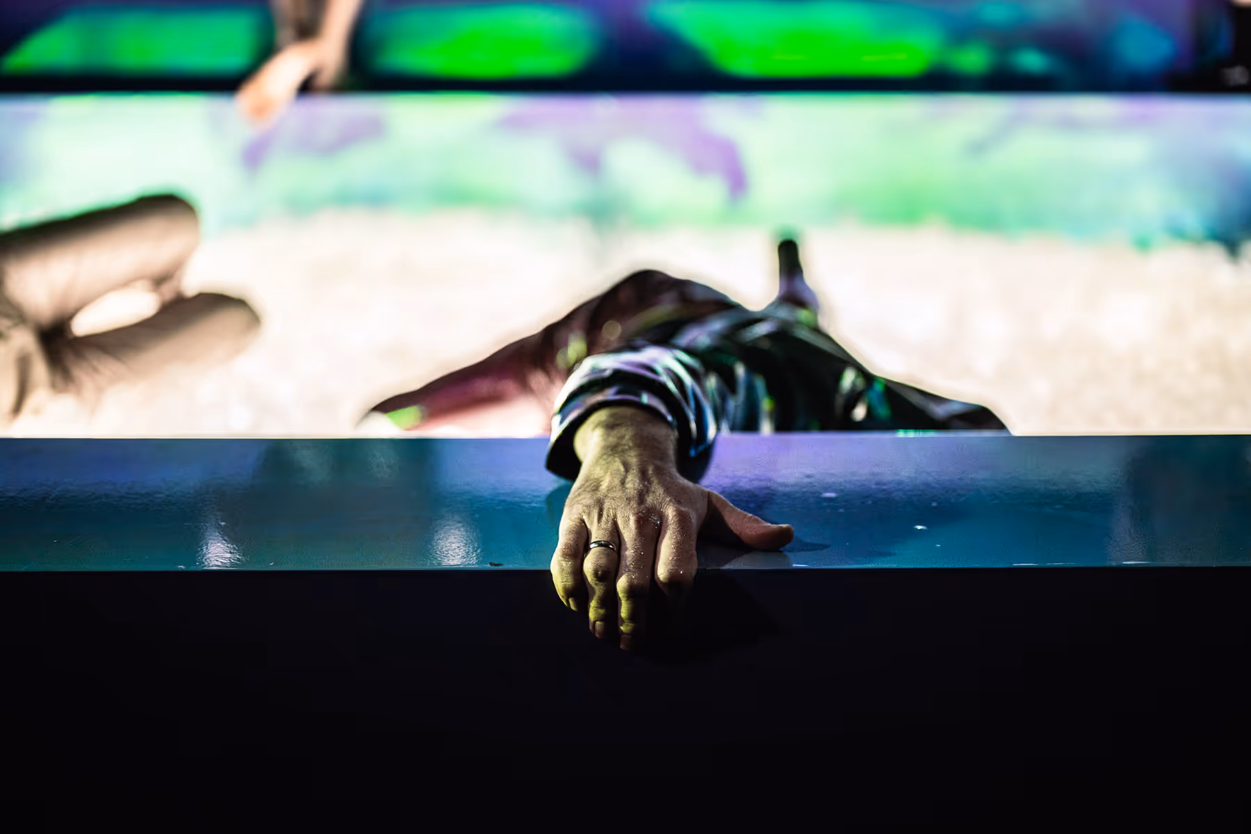 Close-up of a hand with a ring gripping a shiny surface, with a blurred person lying down in the background under colorful lights.