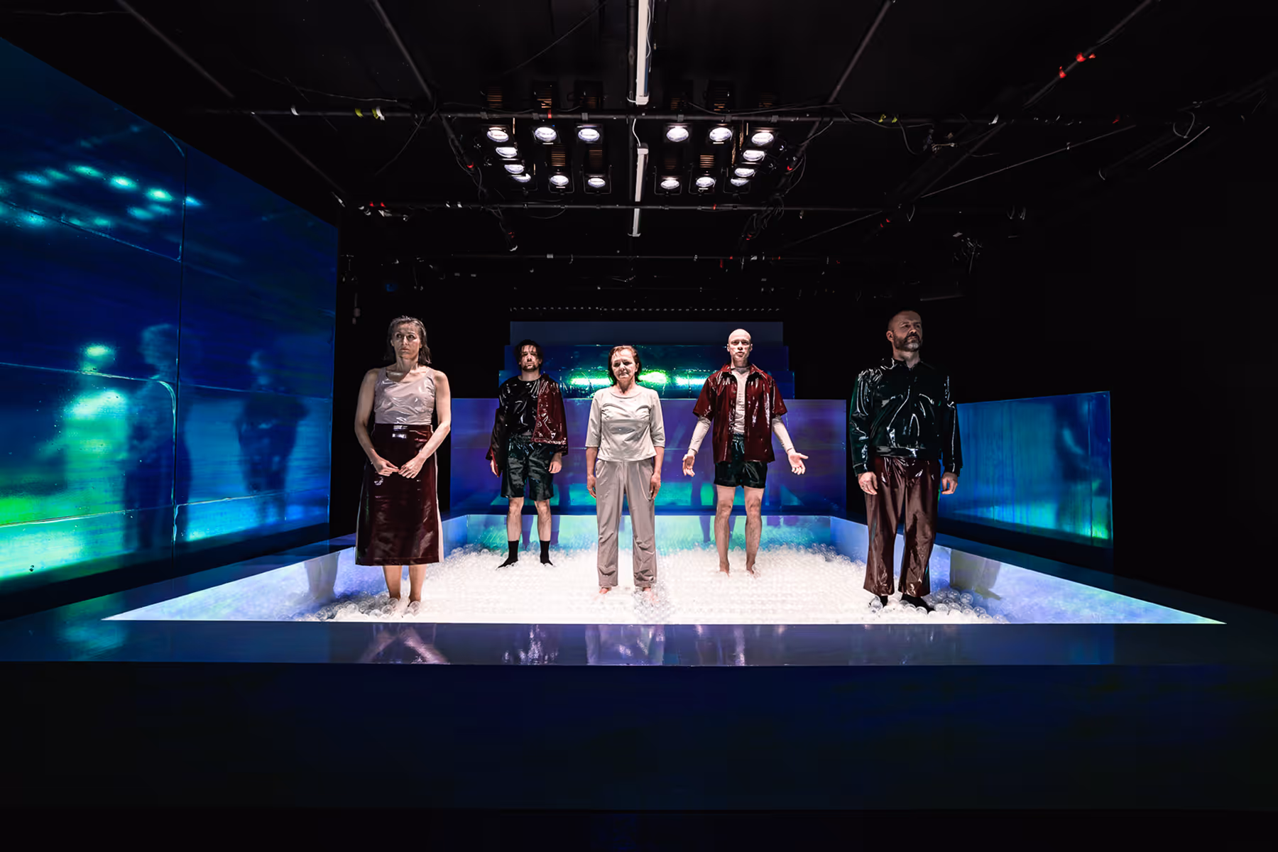 Five people standing on a stage filled with clear plastic balls under bright overhead lights with blue illuminated panels behind them.