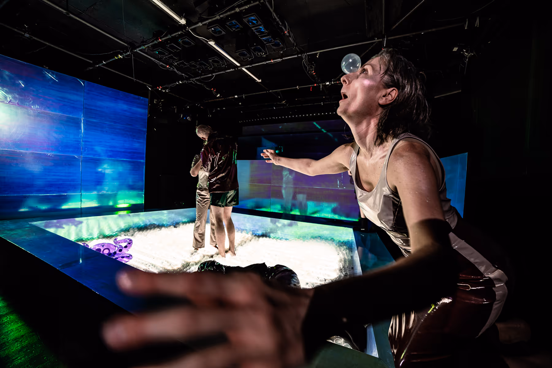 Woman in metallic outfit balancing a bubble on her nose near a shallow pool with two people standing inside, in a dark room with blue lighting.