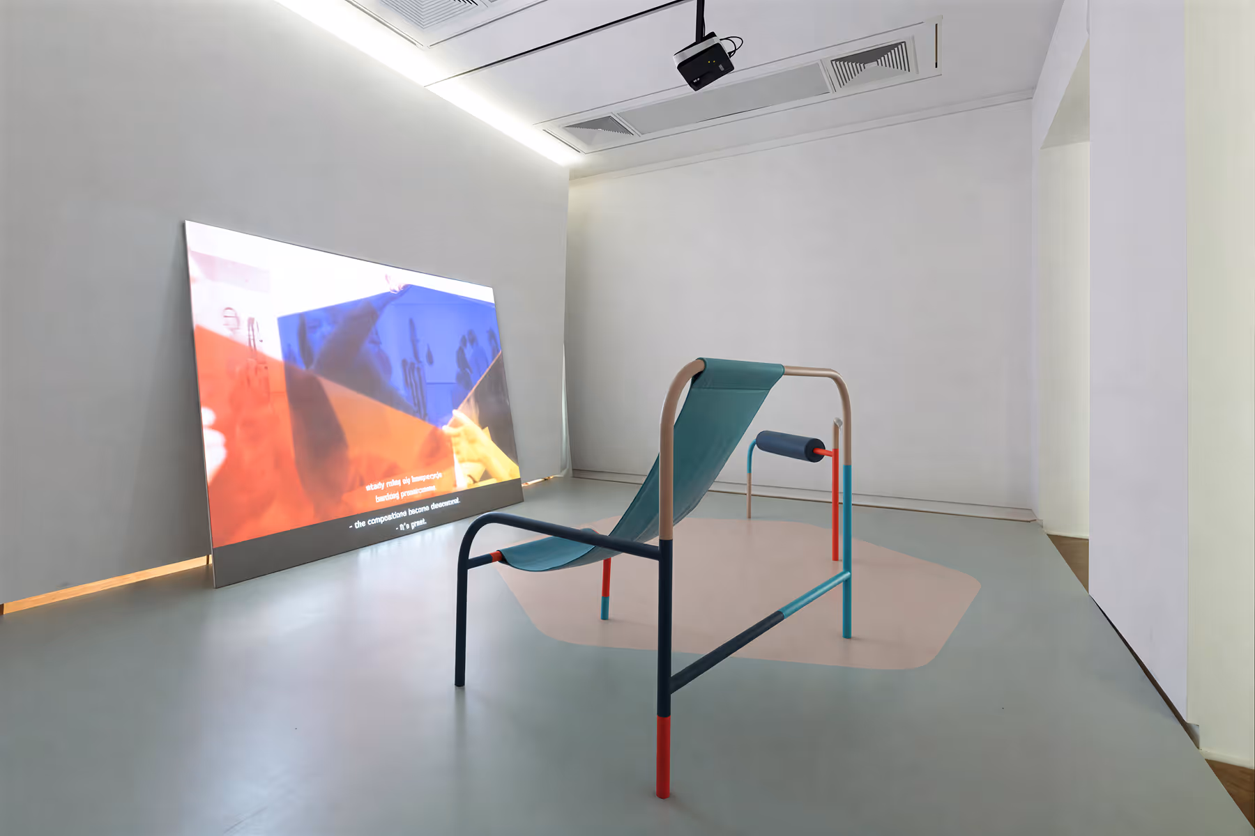 Modern art installation featuring a minimalist chair with colorful legs and a large angled screen projecting abstract visuals in a white gallery space.