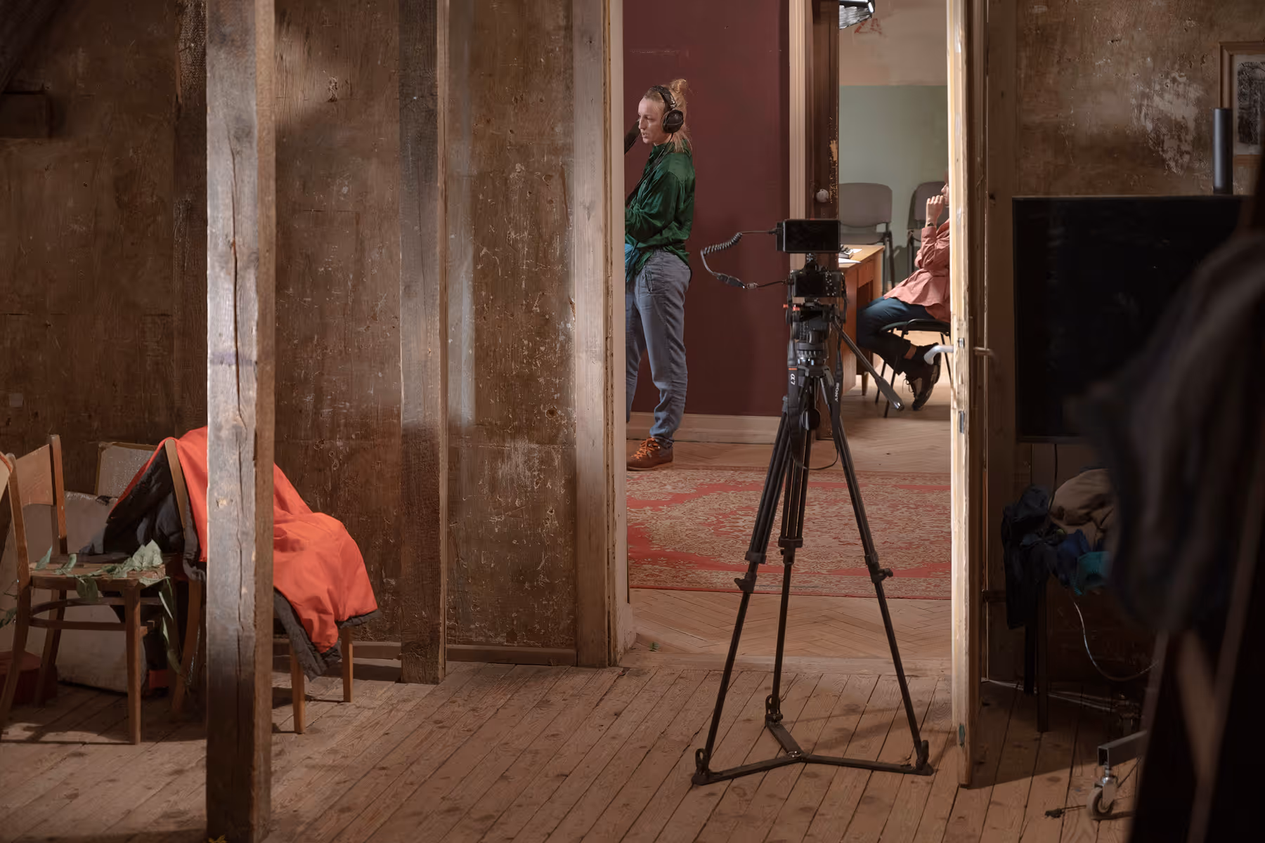 A woman with headphones stands in a doorway next to a camera on a tripod in a rustic room, with a man sitting at a desk in the background.