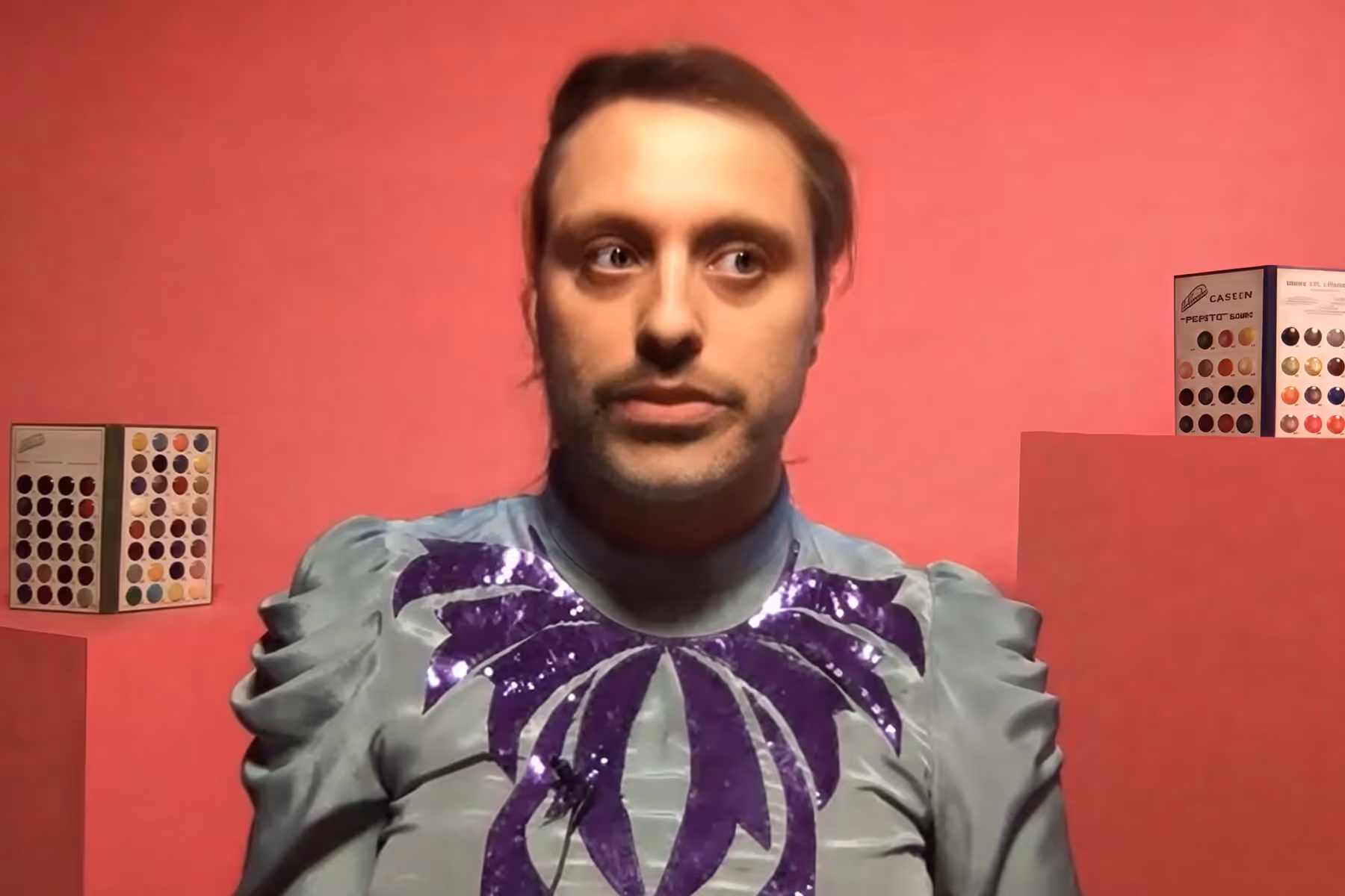 Man with slicked-back hair wearing a light blue shirt with purple sequined design against a pink background.