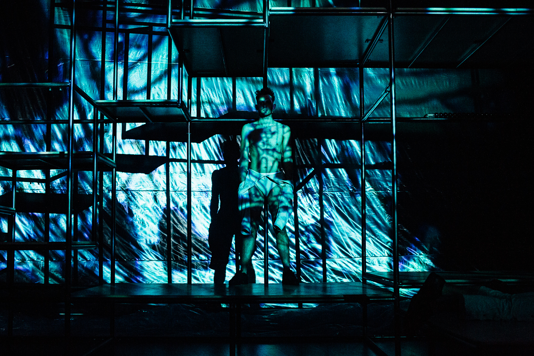Shirtless man standing inside a metal frame structure with blue patterned light projections casting shadows on him and the background.