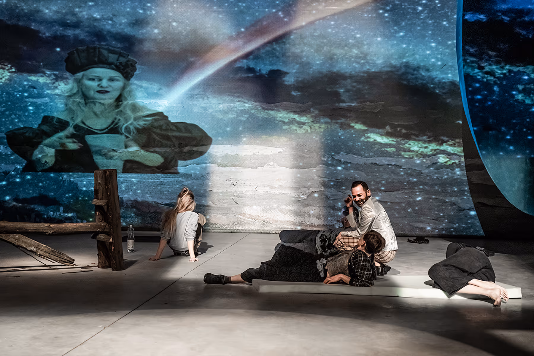 A group of people lounging on a stage floor with a large projection of a woman in historical costume and a cosmic background behind them.