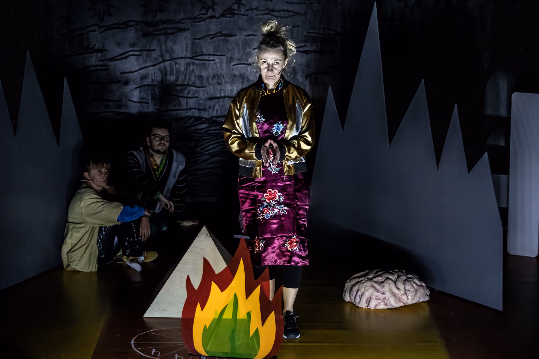 Person in a shiny floral dress and jacket standing in front of a stylized fire prop with two people sitting in the dark background near geometric shapes and a brain model on the floor.