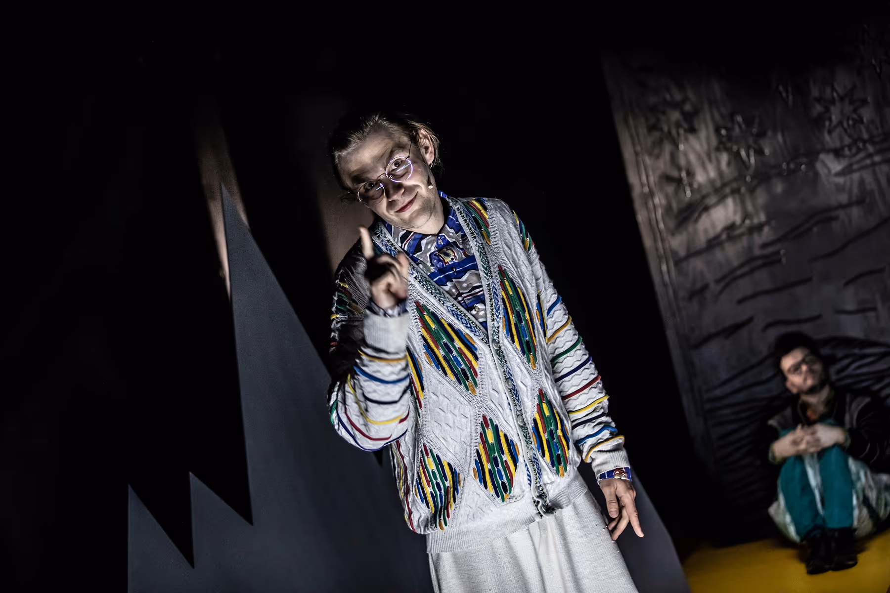 Young man wearing glasses and a colorful patterned sweater sits on stage holding a triangular object, with three other people partially visible behind him.