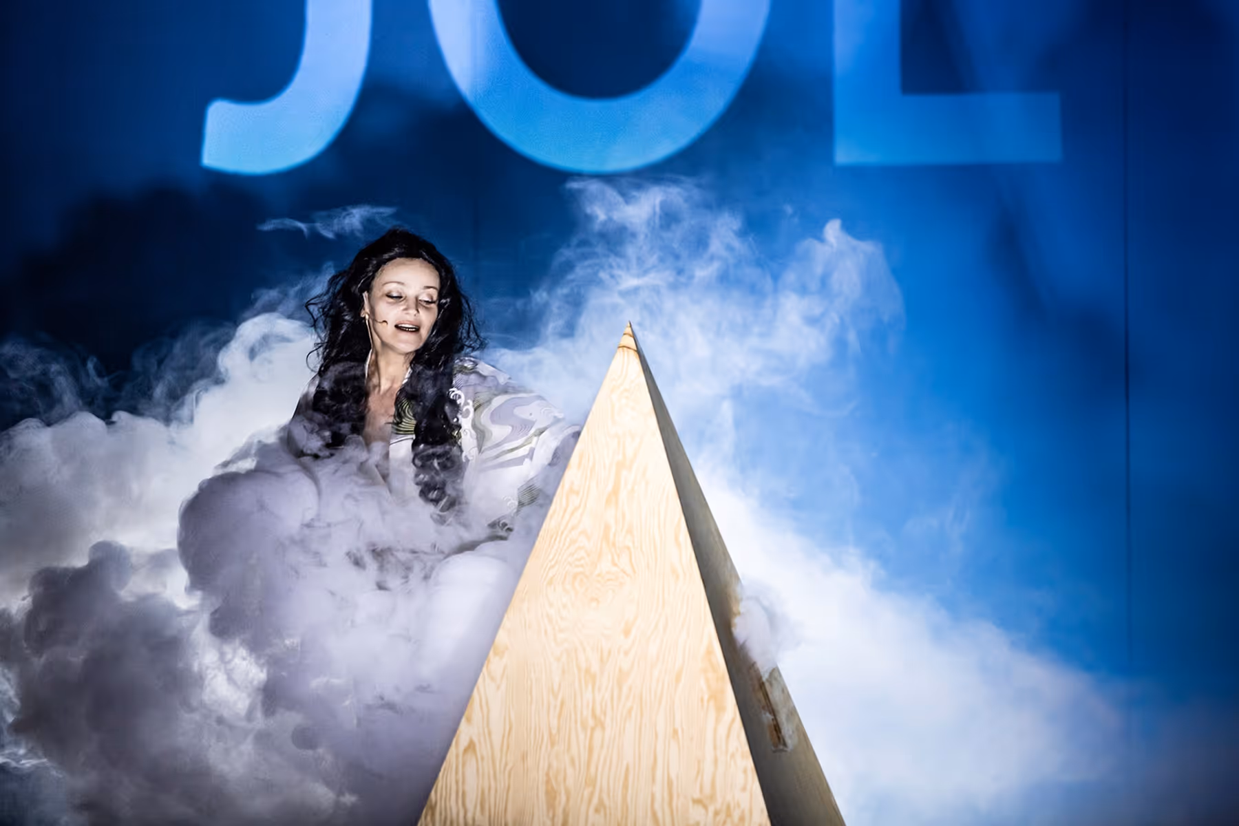 Woman with long dark hair surrounded by smoke standing behind a wooden triangular structure against a blue background.