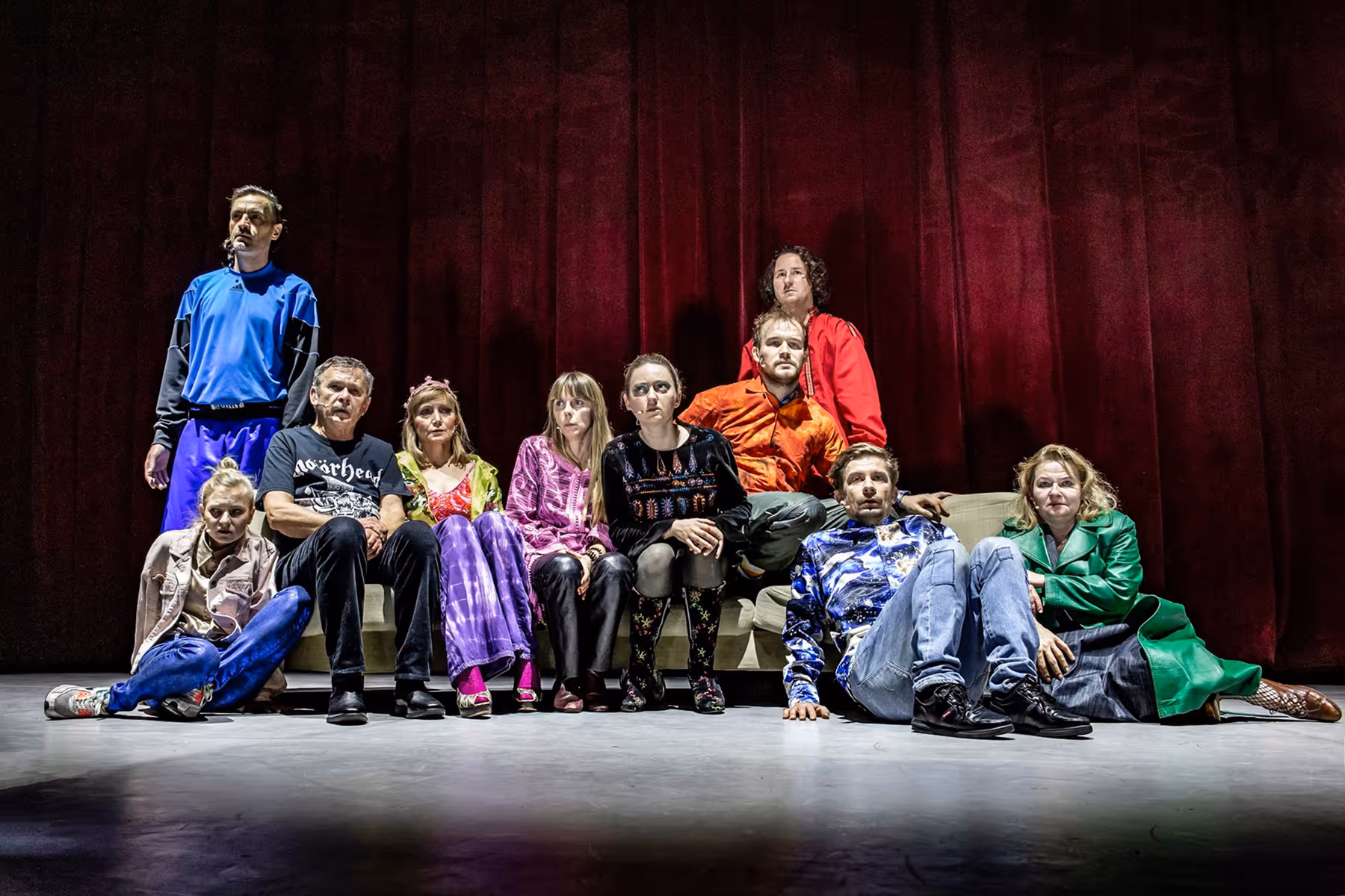 A group of nine people in colorful clothing posed against a dark red curtain background, sitting or kneeling on the floor and couch.