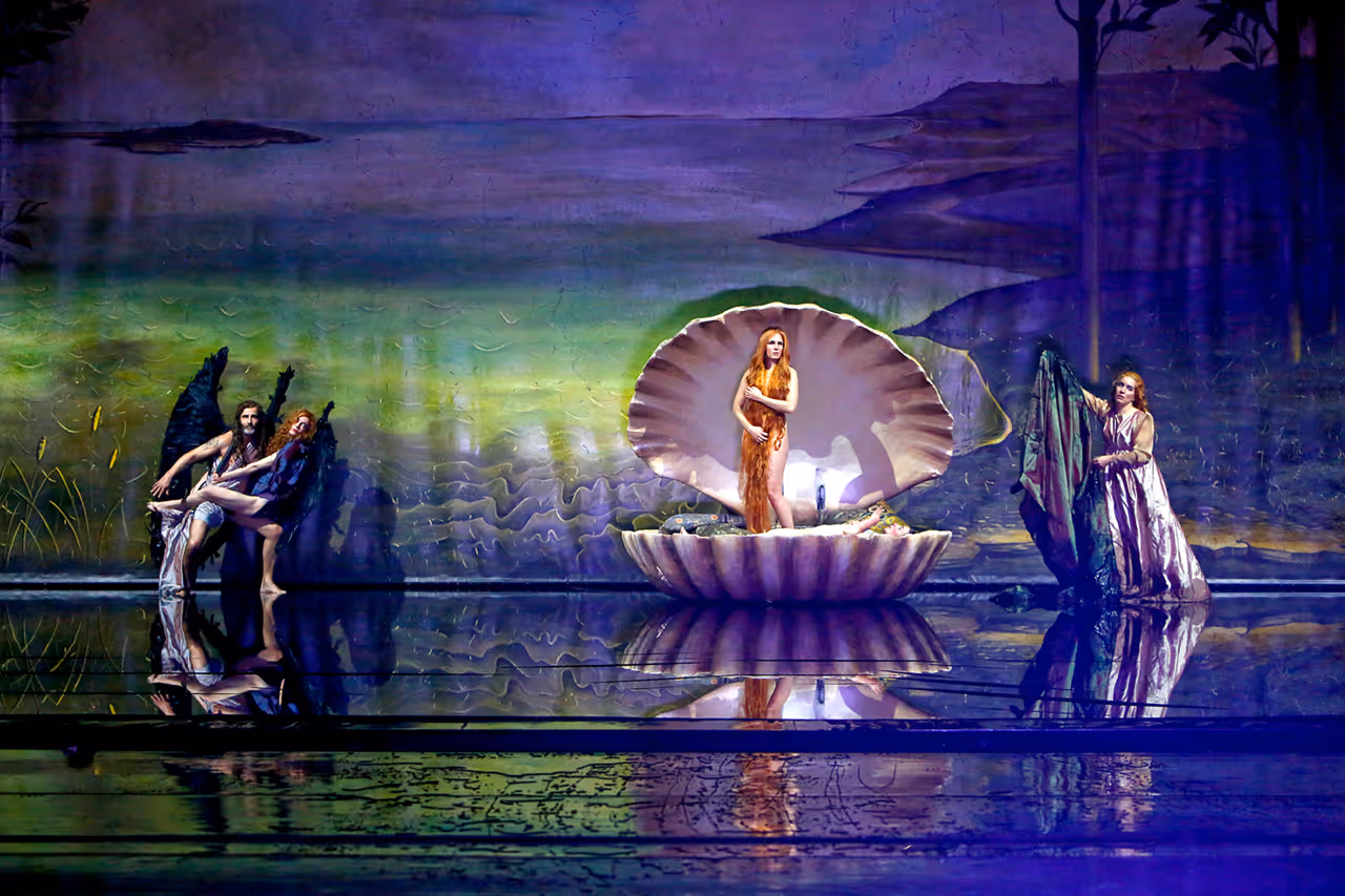 Nude woman sitting in a large green clam shell on stage with a nude man standing nearby, surrounded by green fog and darkness.