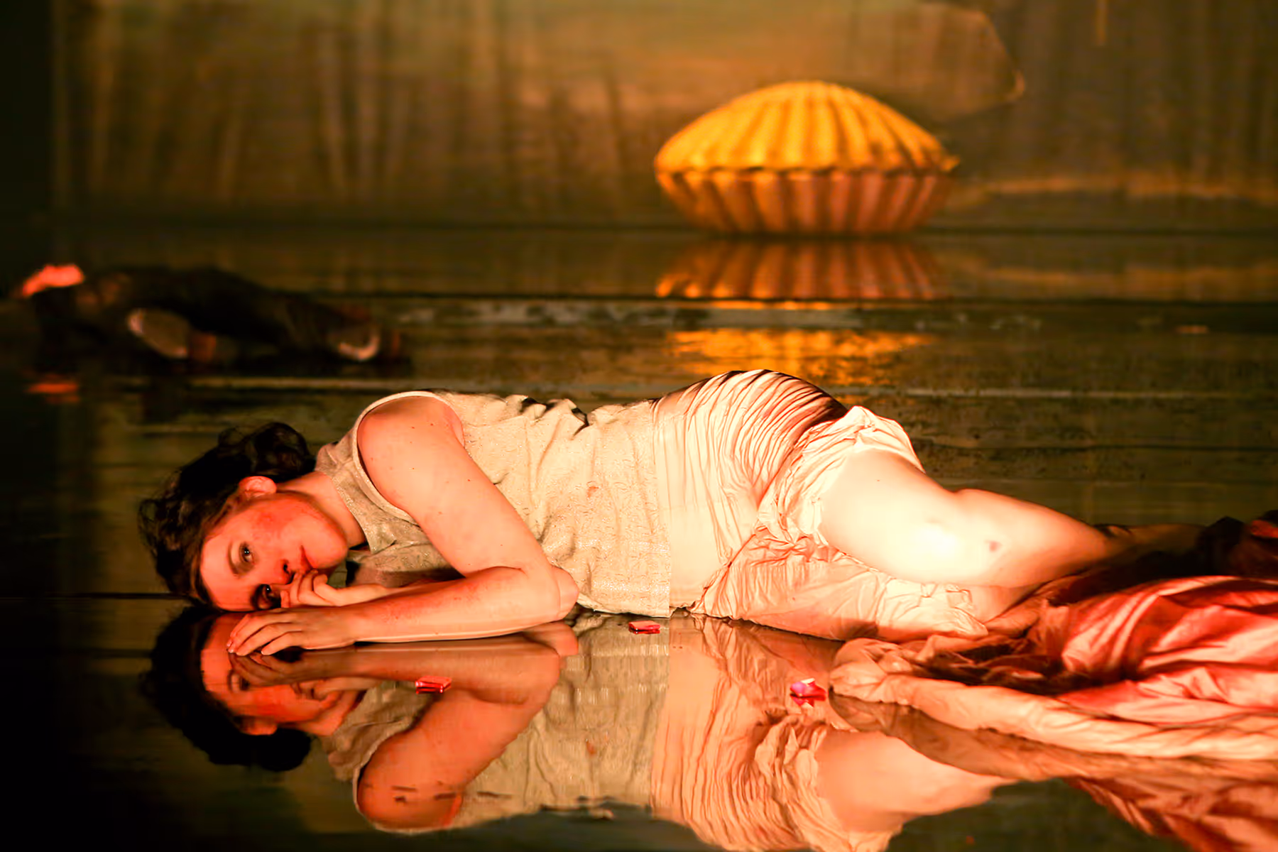 Woman lying on her side on a reflective surface with a golden light and a large round object in the background.