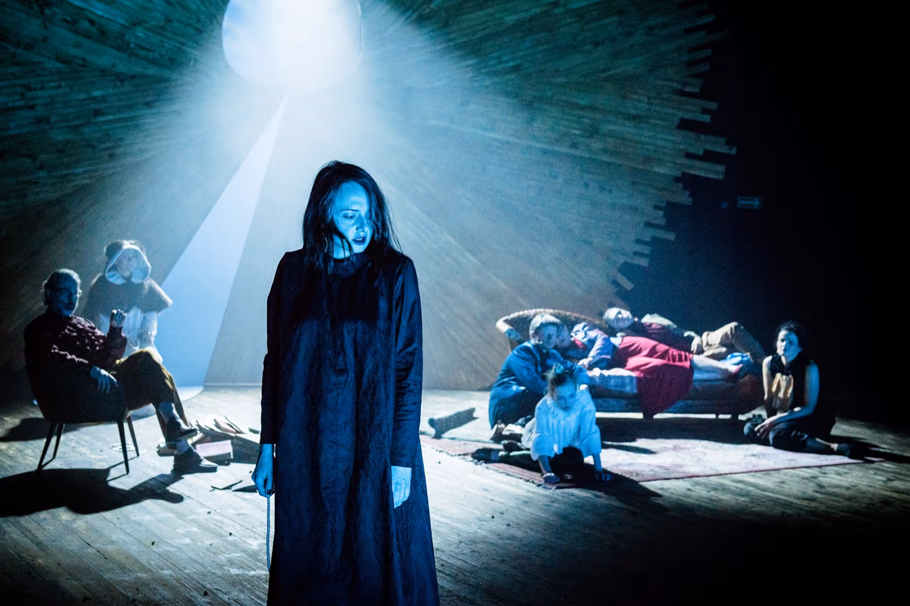 A woman in a long dark dress stands in blue light on a stage while six people sit and lie on chairs and a couch in the background under a spotlight.