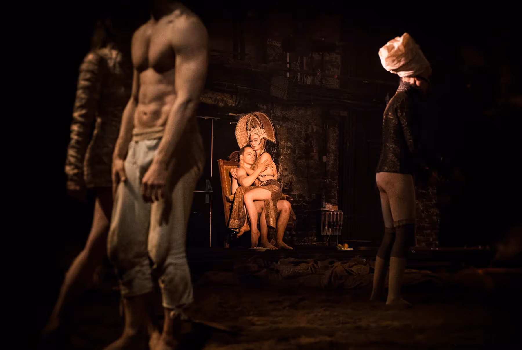 Dimly lit theatrical scene with a shirtless man holding a woman in an ornate headdress sitting on his lap, and three other partially visible figures around them.