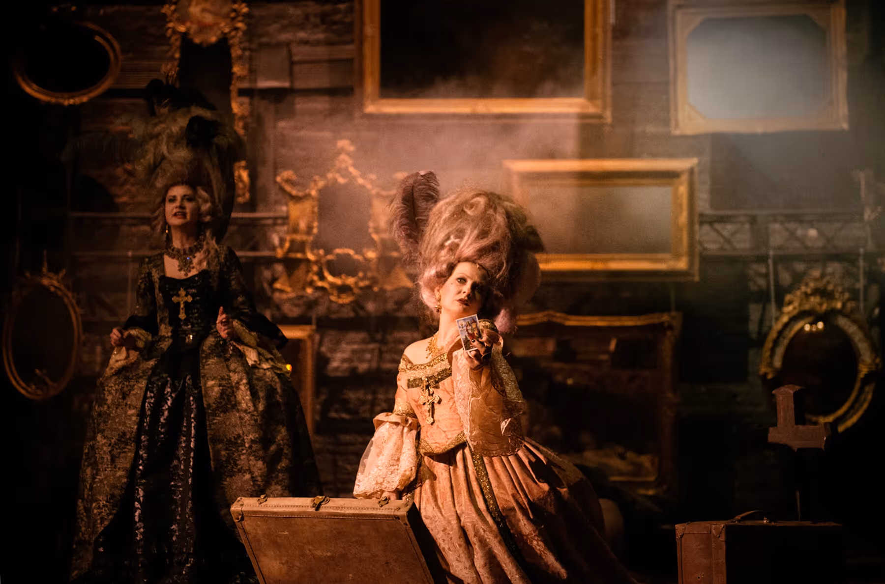 Two women in elaborate 18th-century aristocratic costumes, one holding a tarot card and the other standing beside her in a dimly lit room with empty ornate picture frames on the wall.