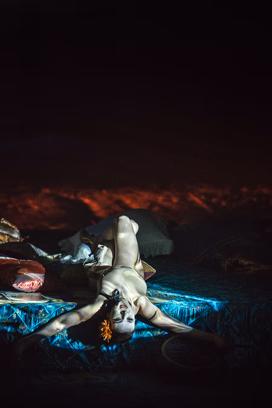 A woman lying on her back on a patterned surface with arms outstretched, wearing a flower in her hair under dramatic lighting.