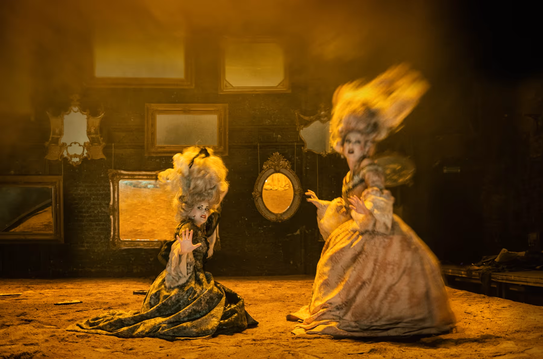 Dimly lit theatrical scene with a shirtless man holding a woman in an ornate headdress sitting on his lap, and three other partially visible figures around them.