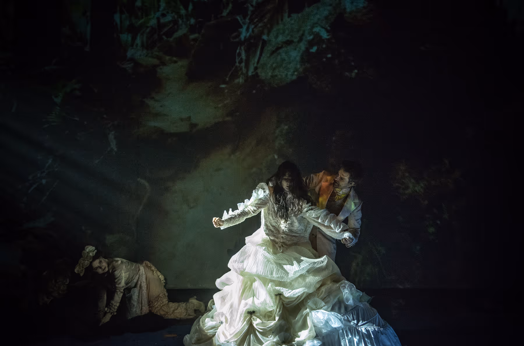Three women in elaborate 18th-century style costumes with powdered wigs, one woman covering the mouth of a doll-like figure.
