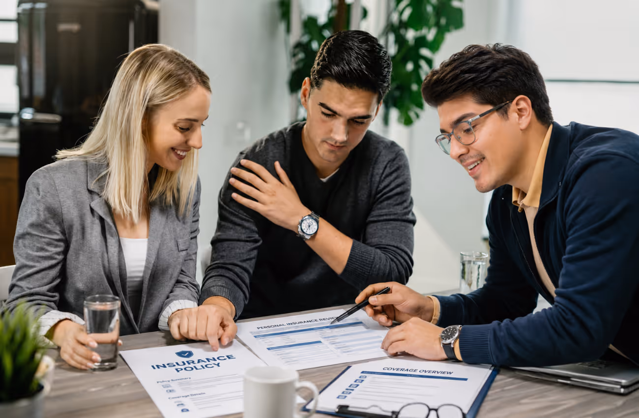 Alonso Contreras reviewing policy documents with a young couple during a consultation in a modern office, explaining coverage options and personal insurance details.