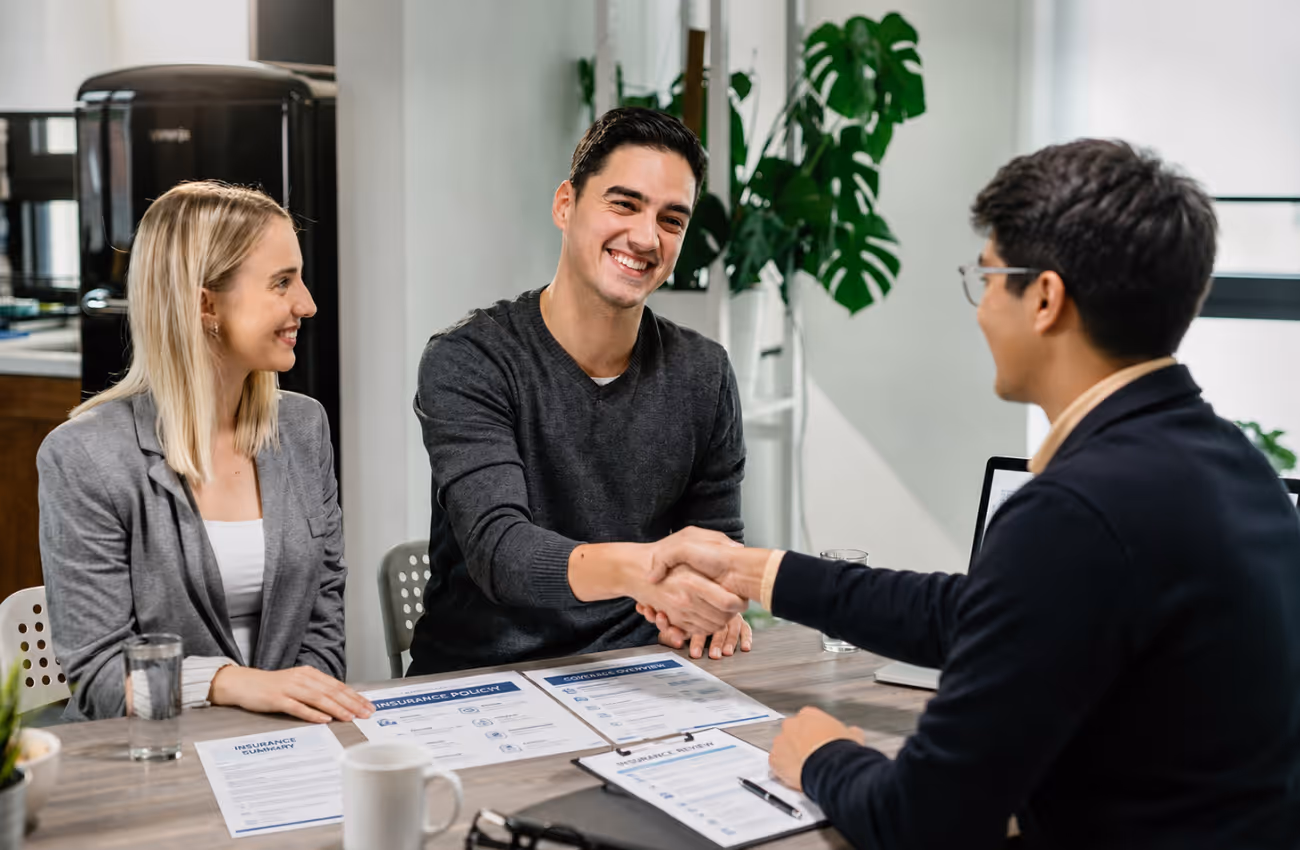 Insurance agent Alonso Contreras shaking hands with a client during a policy review meeting, with insurance documents and coverage forms on the table.