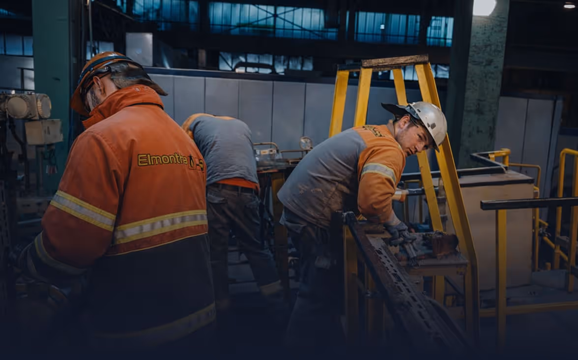 Drei Arbeiter mit Schutzhelmen und Arbeitskleidung in einer Fabrikhalle, die an Maschinen arbeiten.