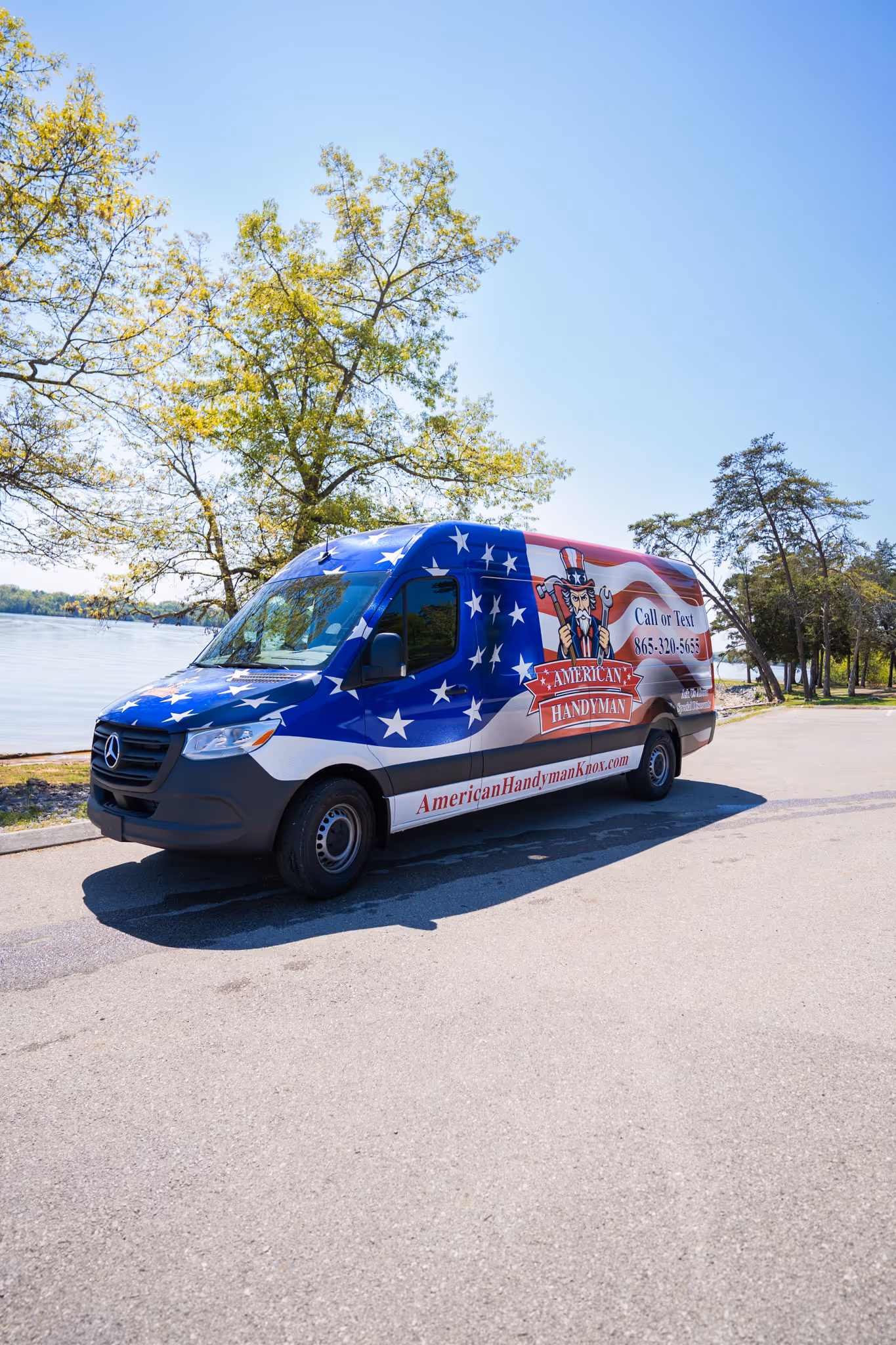 American Handyman service van with patriotic red, white, and blue stars and stripes design parked by a lakeside on a sunny day.