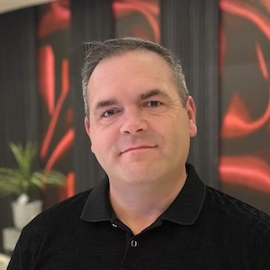 Middle-aged man with short gray hair wearing a black collared shirt, smiling indoors with blurred background.