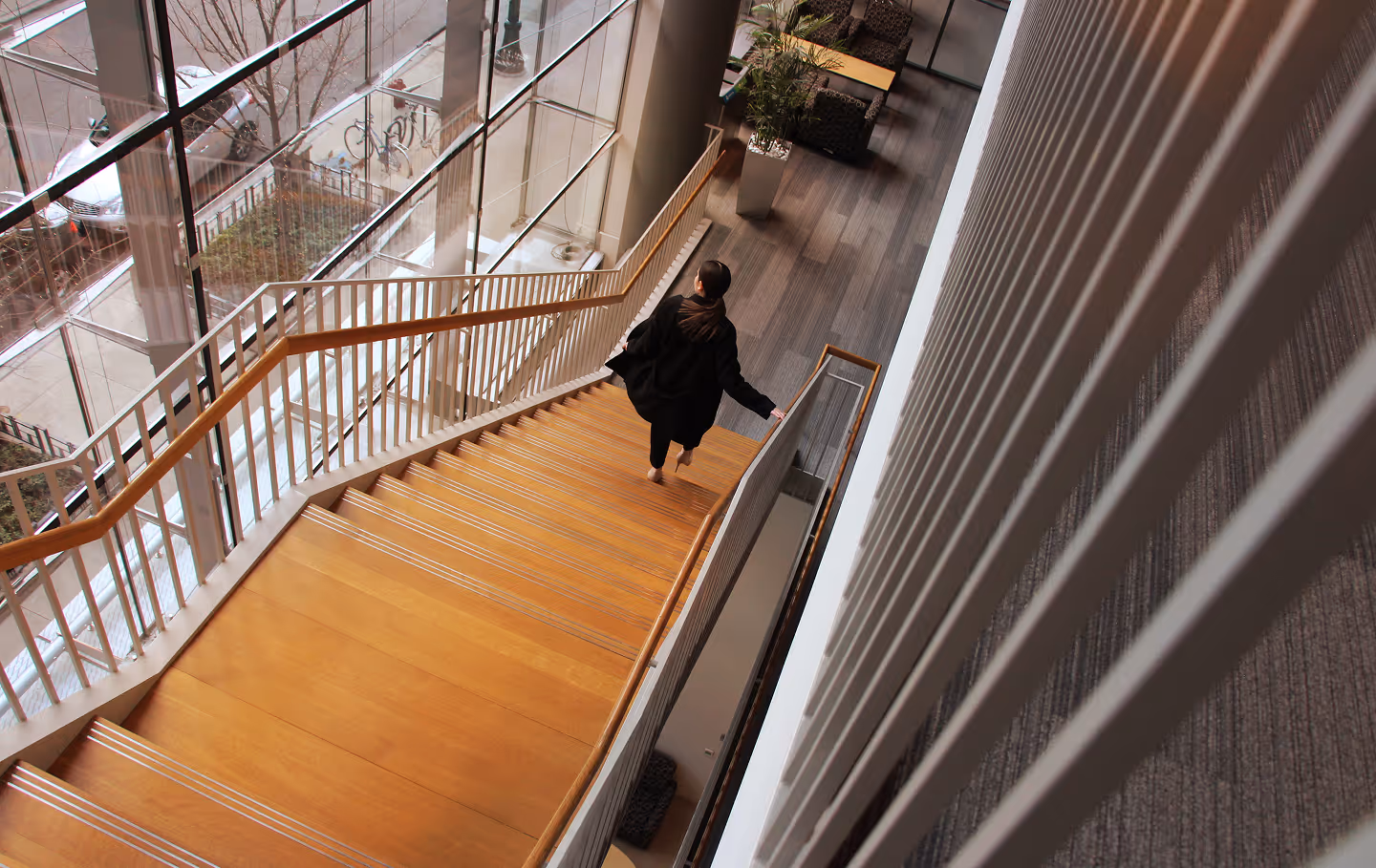 Woman in black coat walking barefoot down wooden stairs inside a building with large windows.