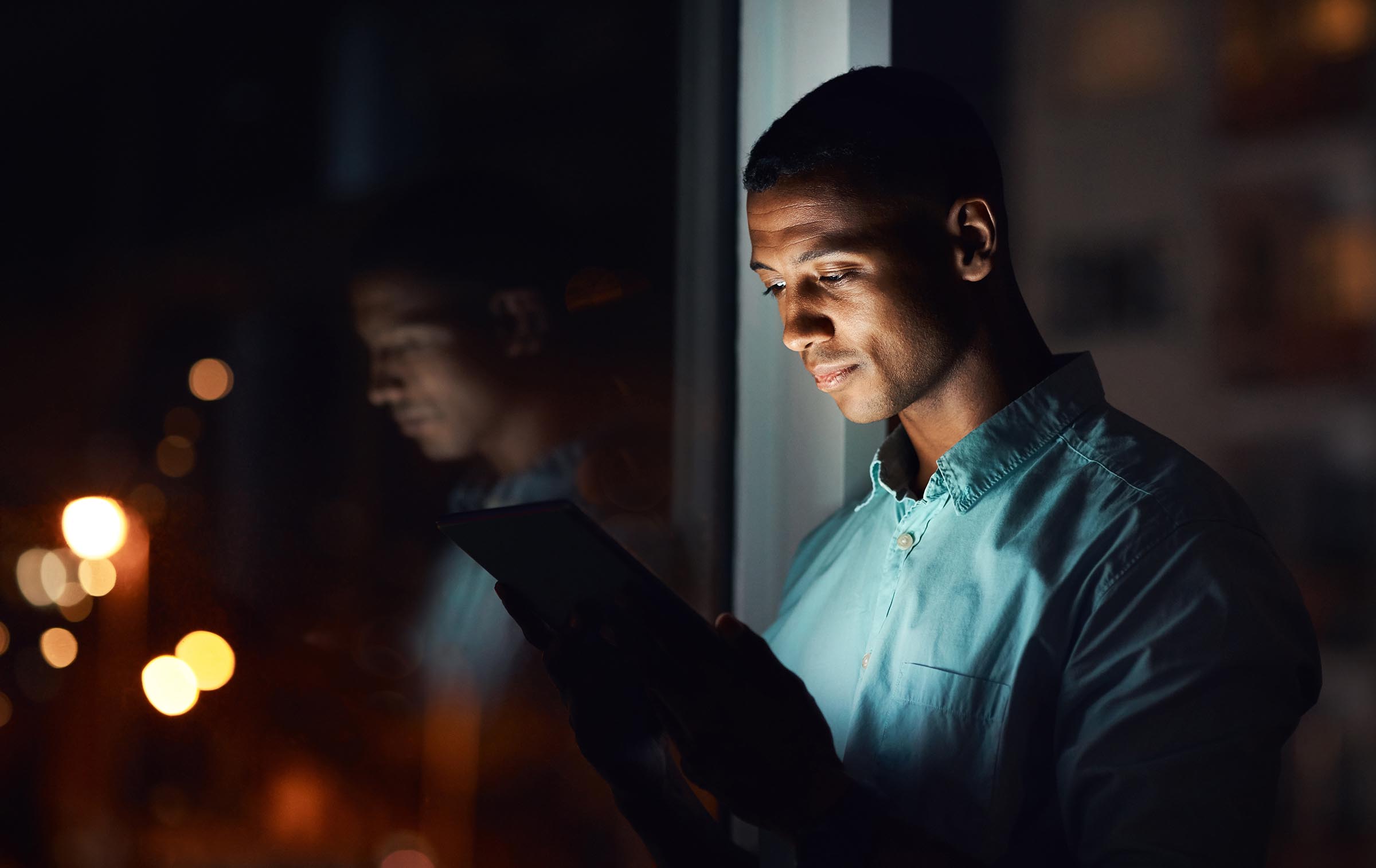 Man standing by window at night reading or using a tablet with light illuminating his face.