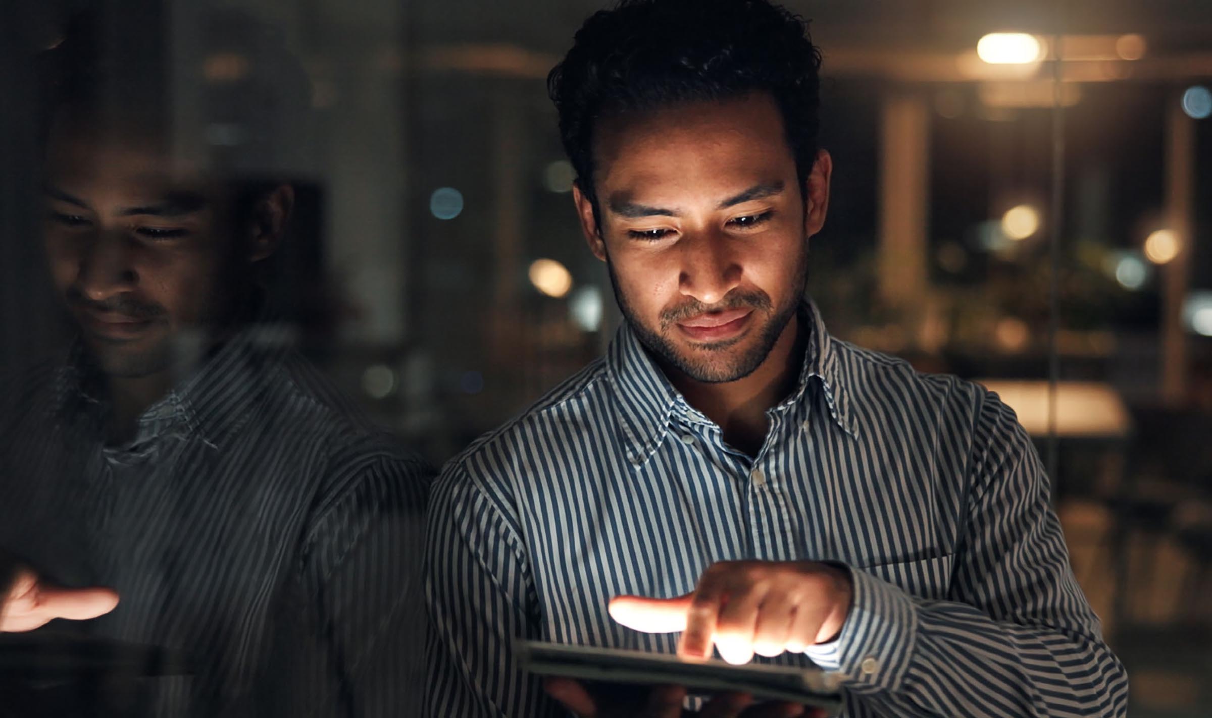 Man in striped shirt using a tablet, illuminated by its light in a dimly lit room at night.