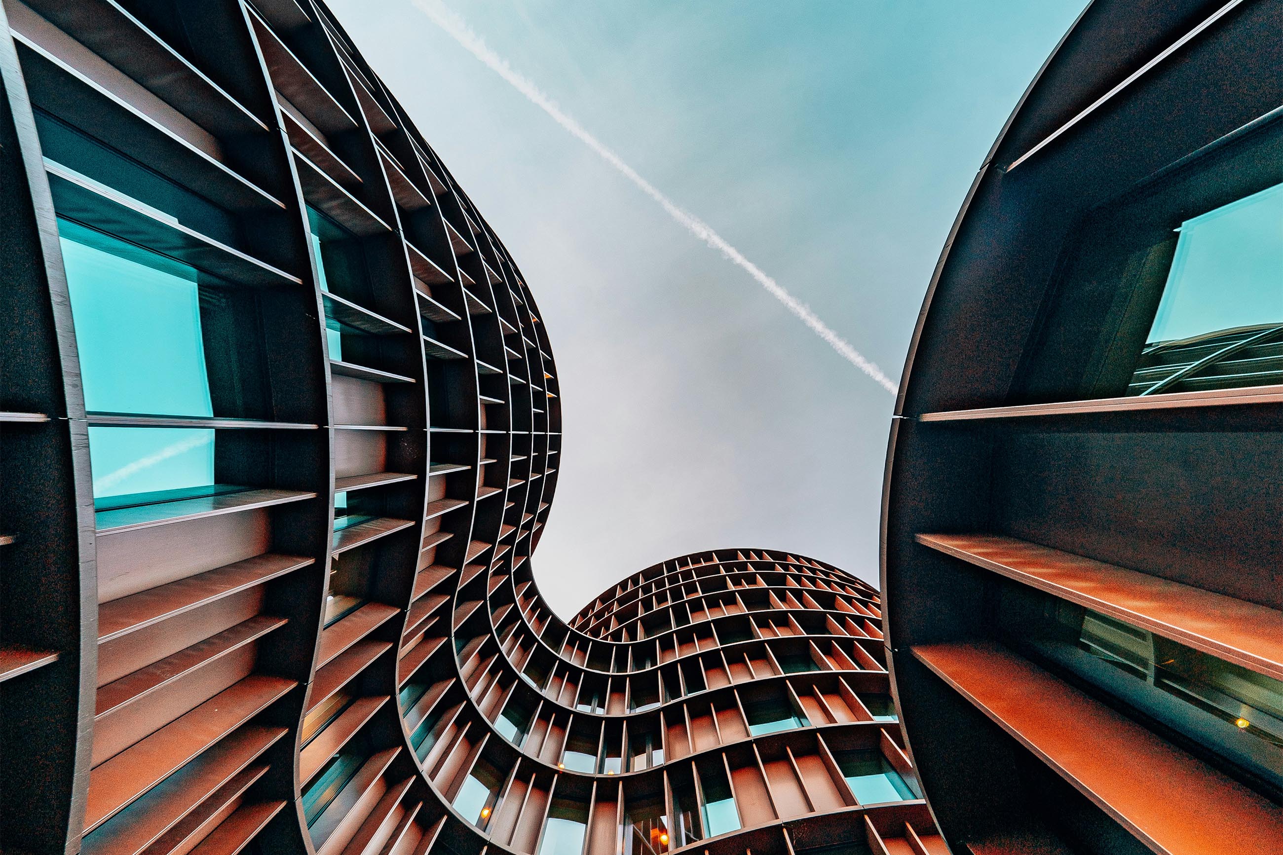 Curved modern building with brown vertical panels and blue windows under a clear sky with a contrail.
