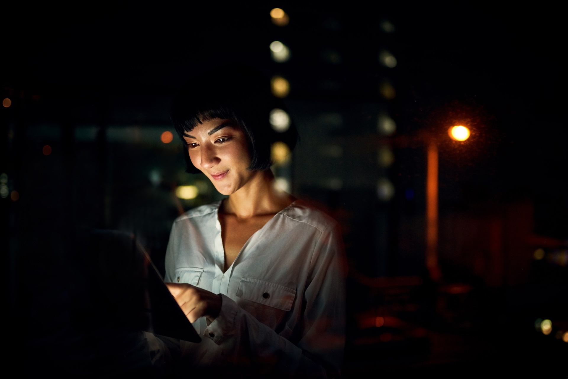 Woman in a white blouse using a tablet at night with city lights in the background.